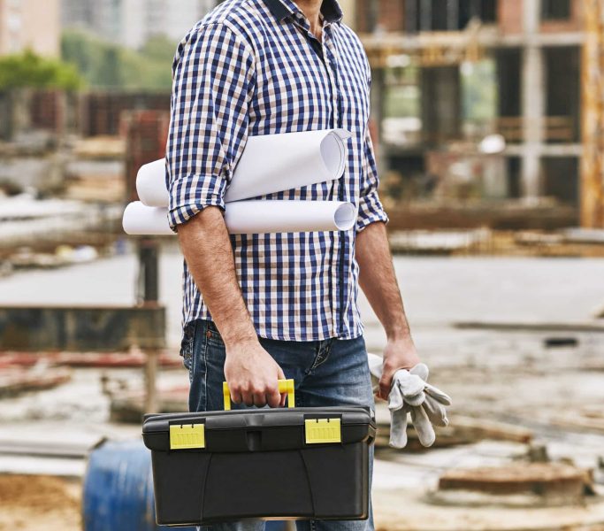 Tout ce dont j'ai besoin pour le travail. Image recadrée d'un constructeur tenant une boîte à outils, des gants de protection et des dessins techniques alors qu'il se tient debout sur un chantier de construction. Concept de construction. Équipement de protection. Concept de construction